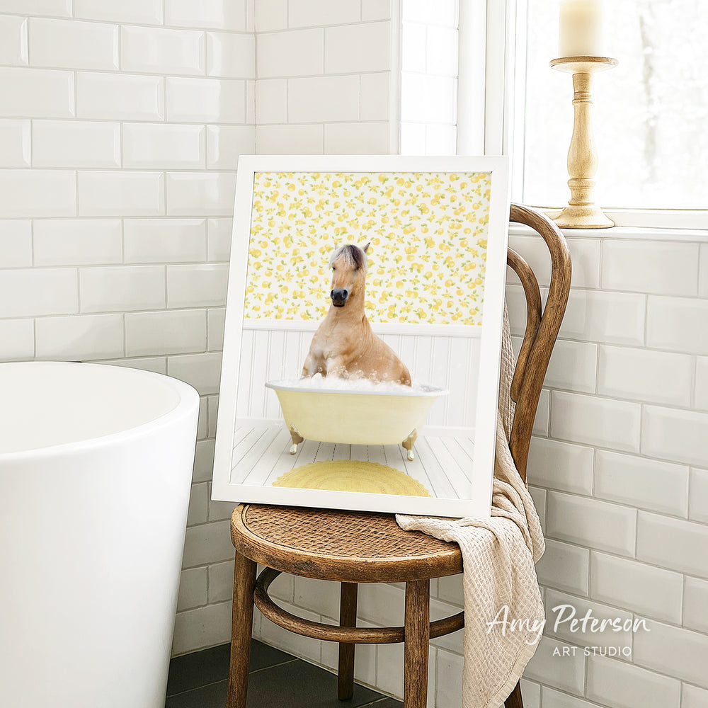 A framed image of a brown horse sitting in a bathtub, surrounded by yellow flowers, is displayed on a wooden chair in a bathroom setting.