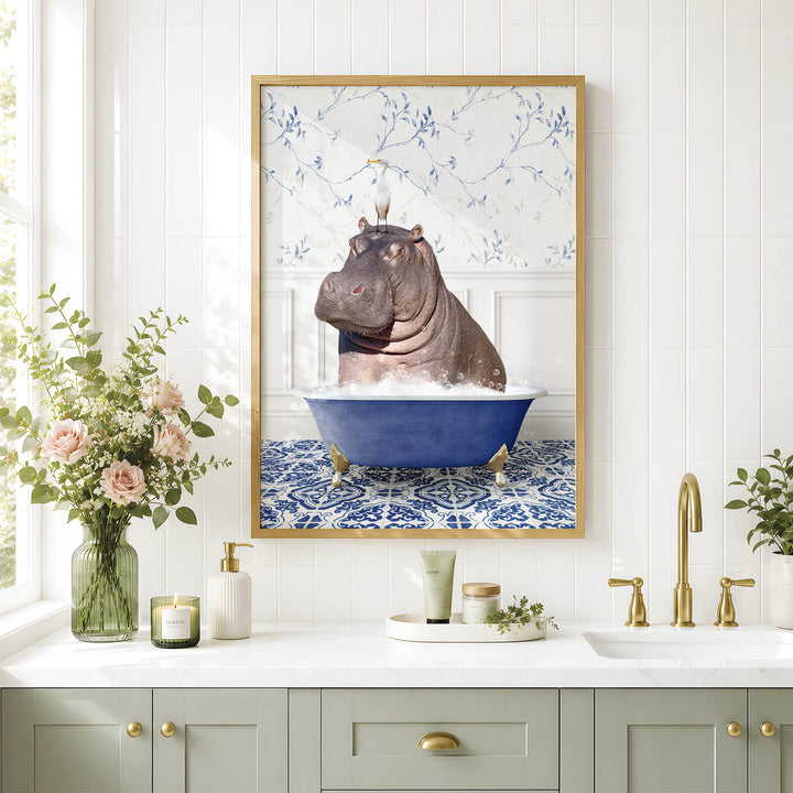 A framed image of a hippopotamus sitting in a blue bathtub, surrounded by flowers and a decorative vase, is displayed on a white tiled wall above a bathroom sink.
