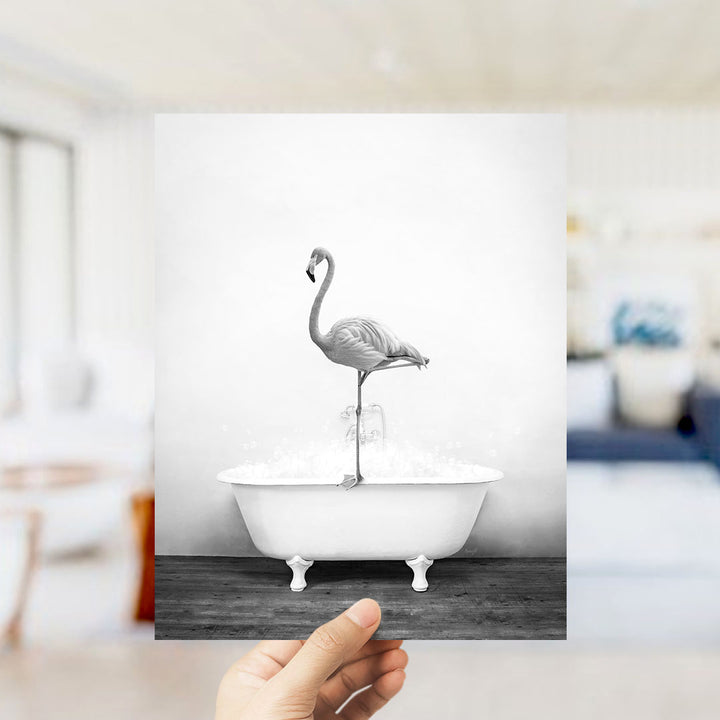 A hand holds up a black and white photograph of a flamingo standing on the edge of a bathtub filled with water, creating a whimsical and surreal scene.
