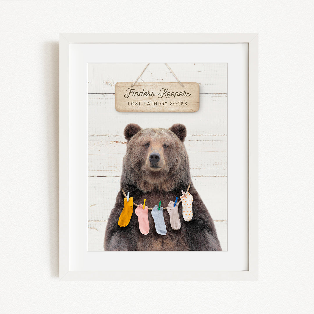 A framed image of a brown bear wearing colorful socks hanging on a clothesline, with a sign above that says "Finders Keepers Lost Laundry Socks".