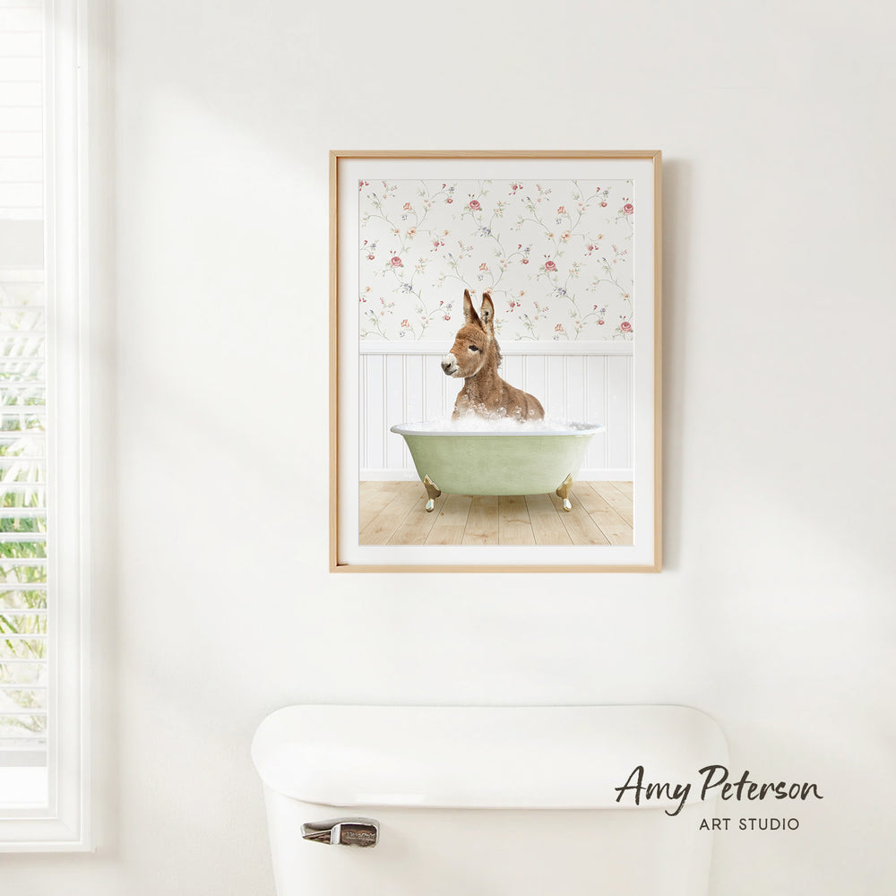 A framed print of a donkey sitting in a bathtub is hanging on a white wall in a bathroom.