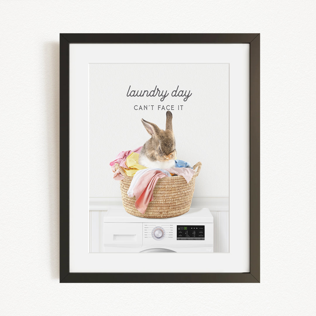 A framed image depicting a rabbit sitting in a woven laundry basket filled with clothes, with the text "laundry day can't face it" above the image.