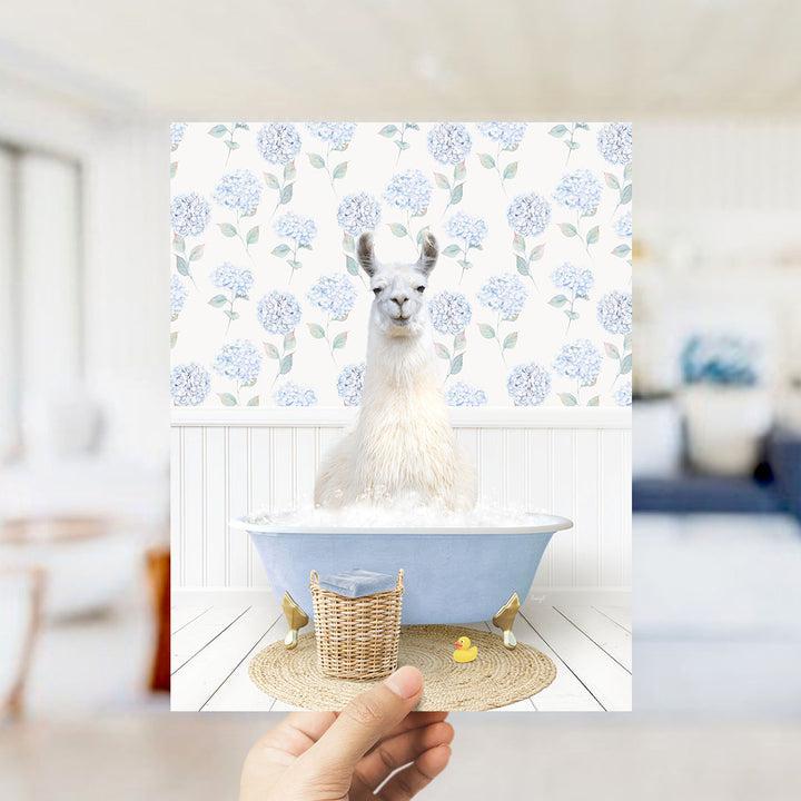 A white llama sitting in a blue bathtub filled with bubbles, surrounded by a wicker basket and a rubber duck.