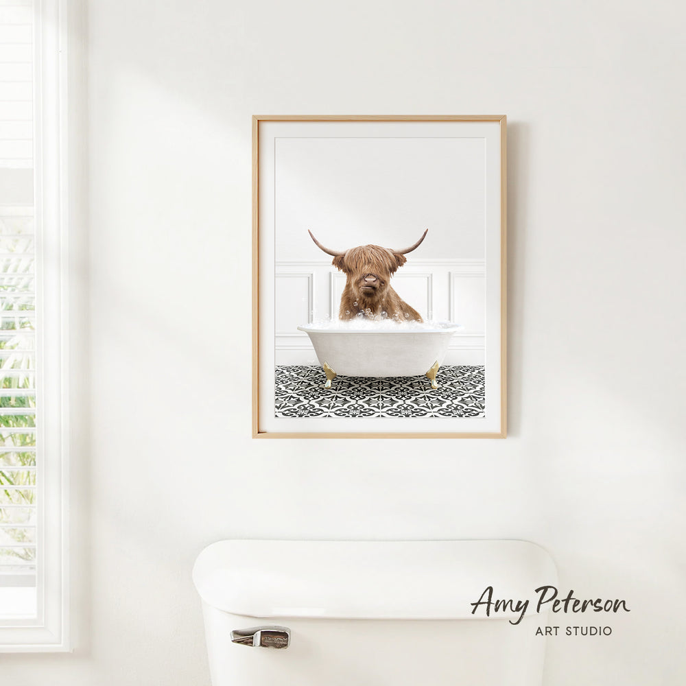 A framed image of a Highland cow sitting in a bathtub, with a patterned tile floor beneath it, is hanging on a white wall.