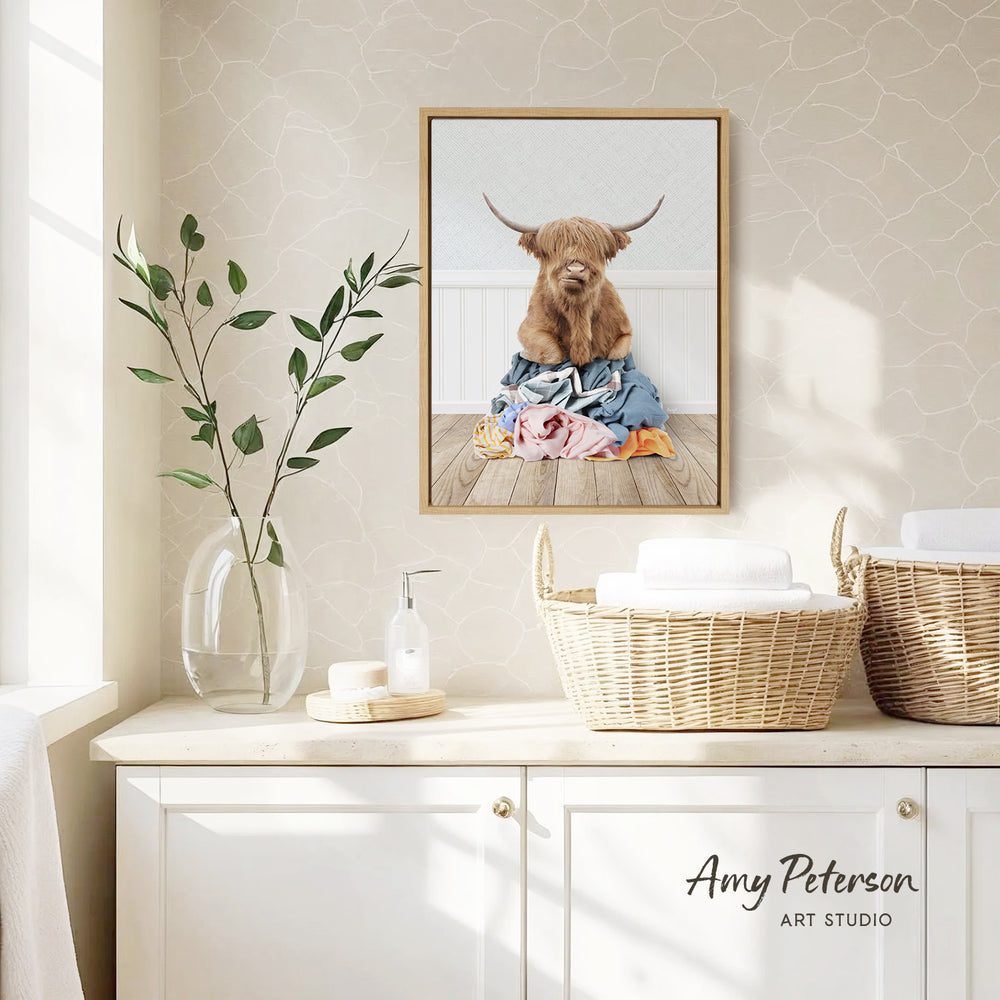 a framed photograph of a Highland cow sitting on a pile of clothes, hanging on a wall above a white cabinet with baskets and a vase of leaves.
