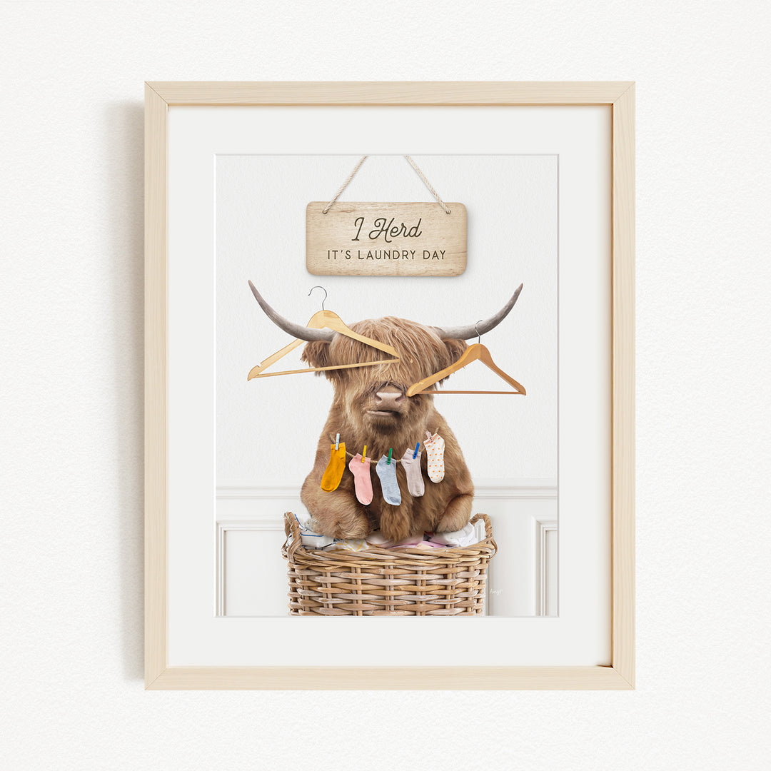 A framed image depicting a brown cow with horns wearing clothes and socks, sitting in a wicker basket with clothes hanging on it.