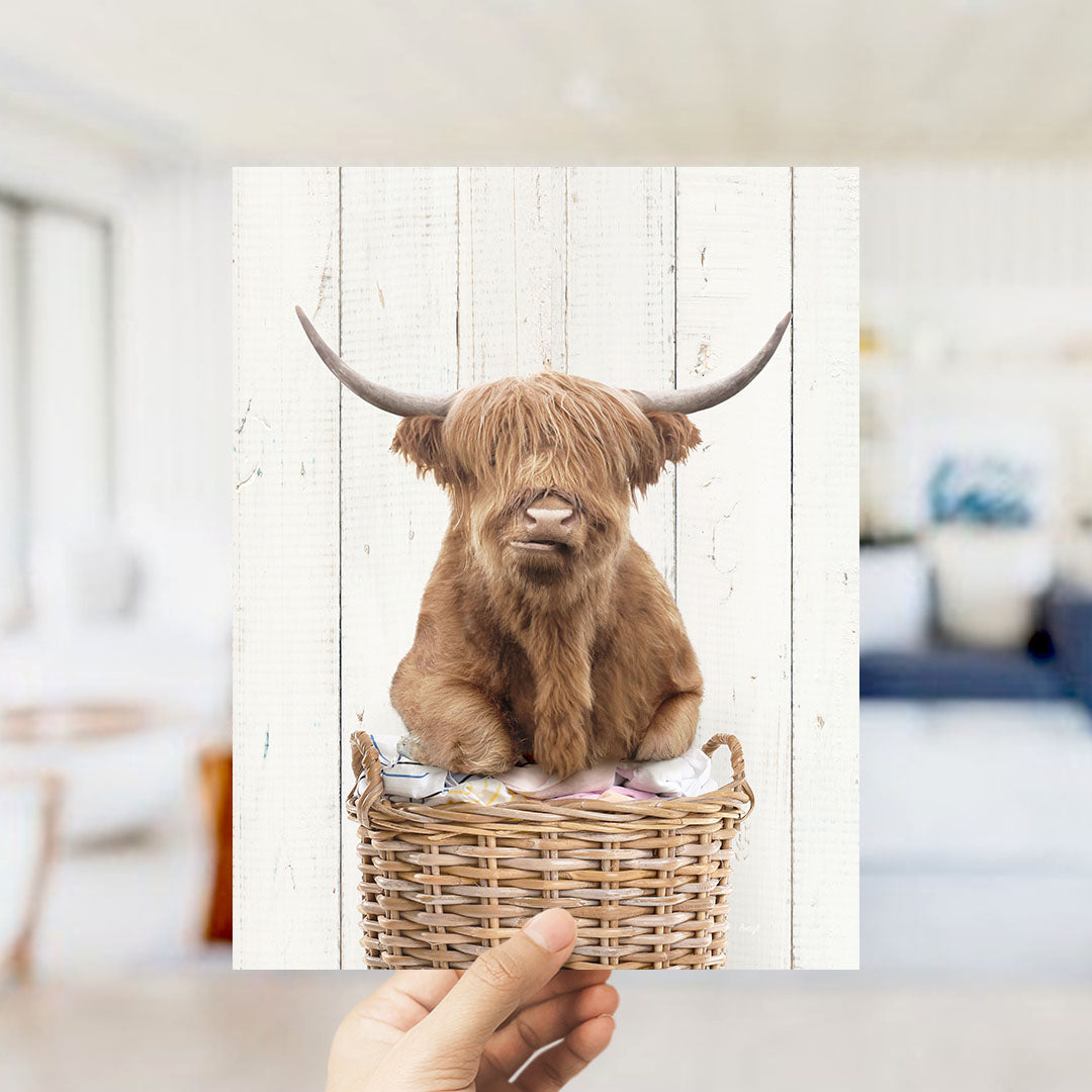 A brown Highland cow with large curved horns is sitting in a wicker basket against a white wooden background.