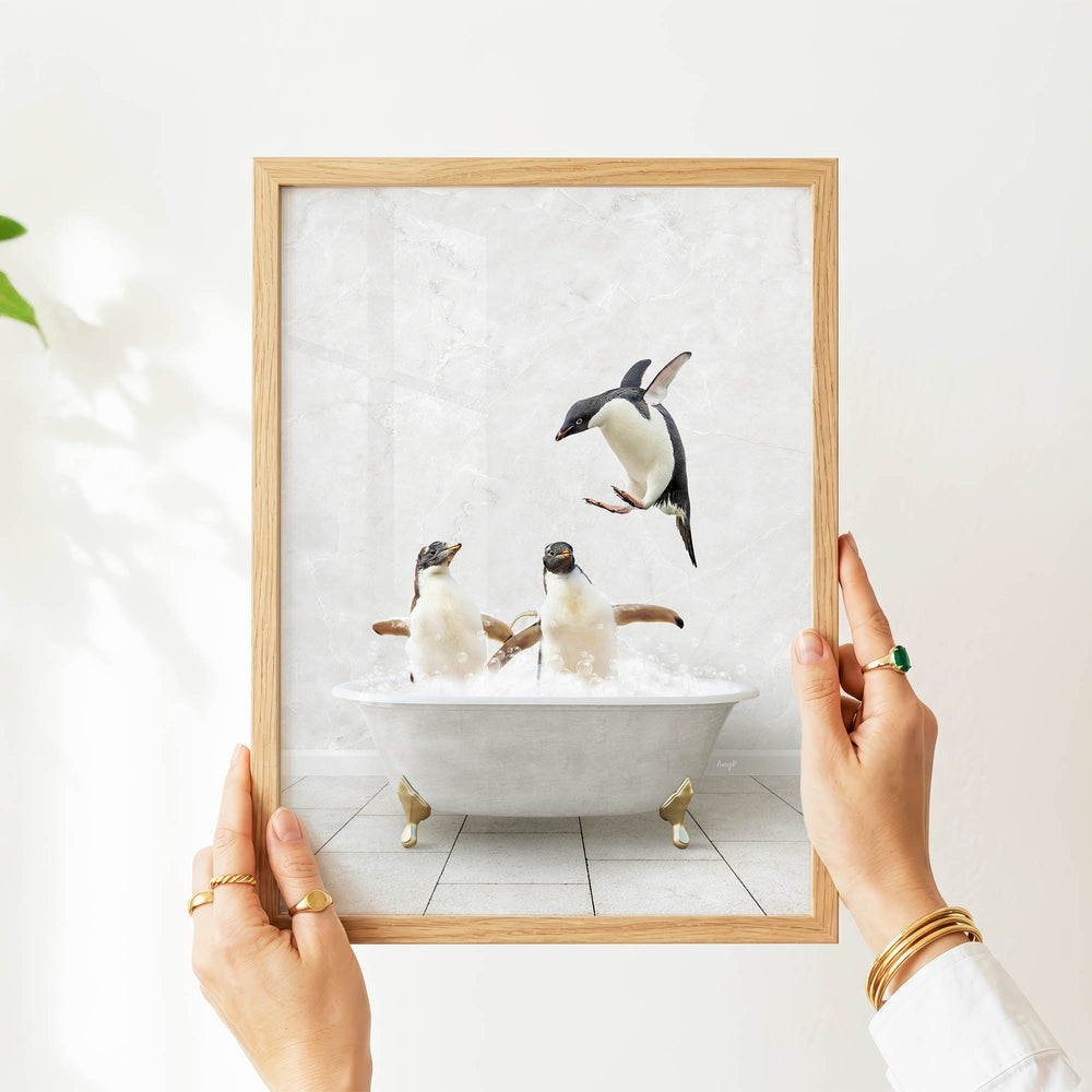 A person's hands are holding a framed picture of two penguins sitting in a bathtub, with one penguin flying out of the tub.