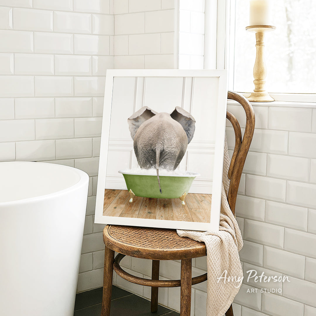 A framed image of an elephant sitting in a green bathtub, with a towel draped over the back of a wooden chair in a bathroom setting.