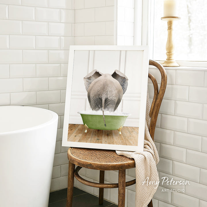 A framed image of an elephant sitting in a green bathtub, with a towel draped over the back of a wooden chair in a bathroom setting.