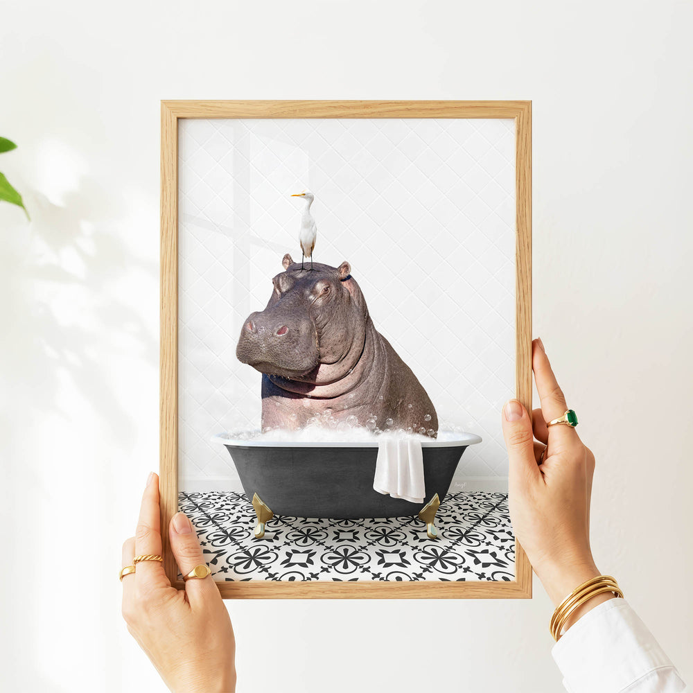A person's hands are holding a framed picture of a hippopotamus sitting in a bathtub, with a bird perched on its head.