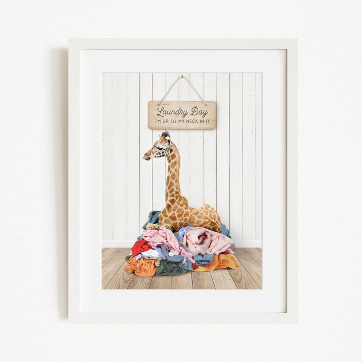 A giraffe is sitting on a pile of clothes in front of a sign that says "Laundry Day".