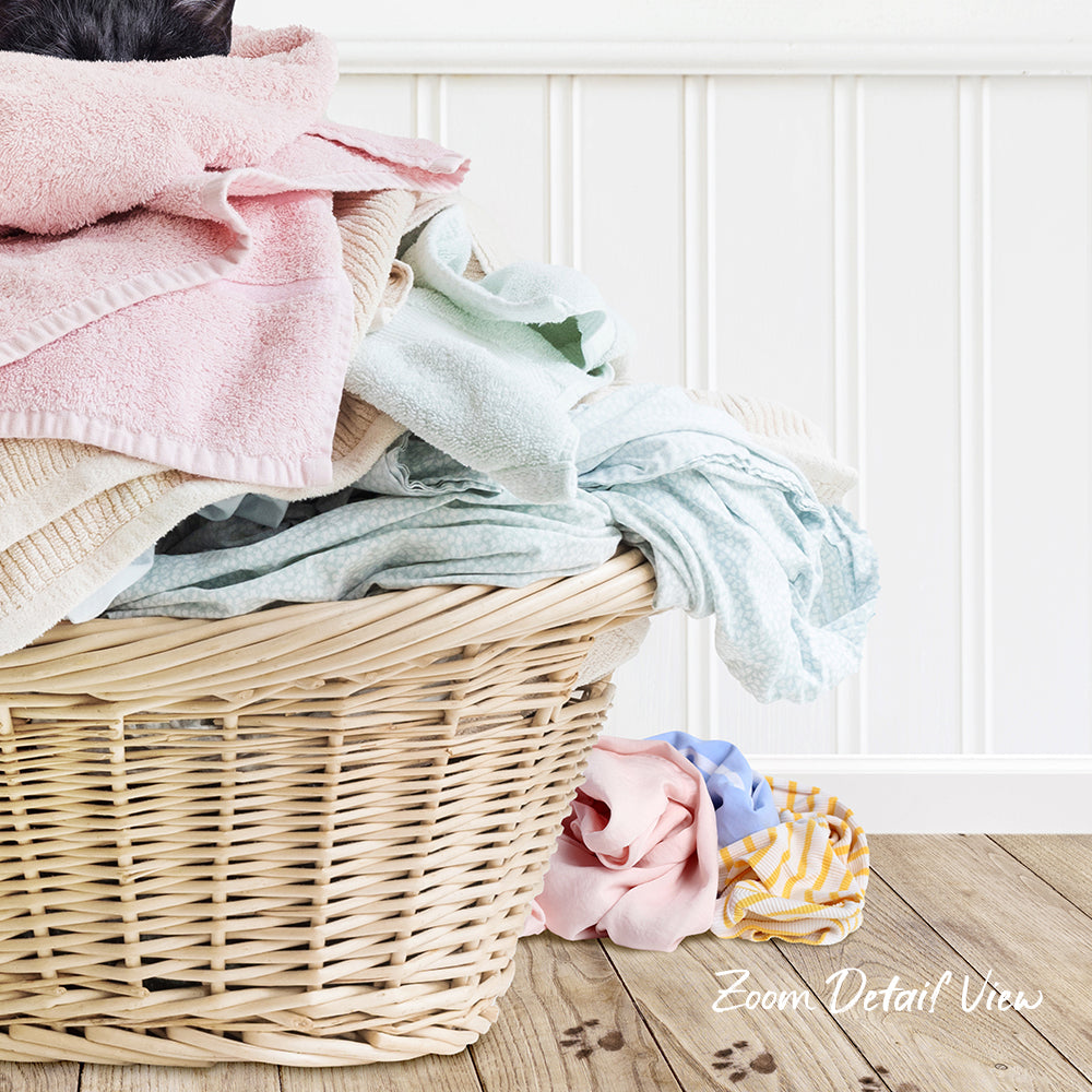 A wicker laundry basket filled with various folded clothes, including a pink towel, sits on a wooden floor against a white wall.
