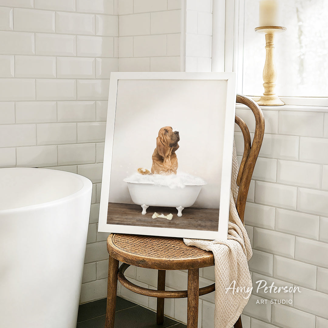 A framed photograph of a dog sitting in a bathtub, with a towel draped over the back of a wooden chair in a bathroom setting.