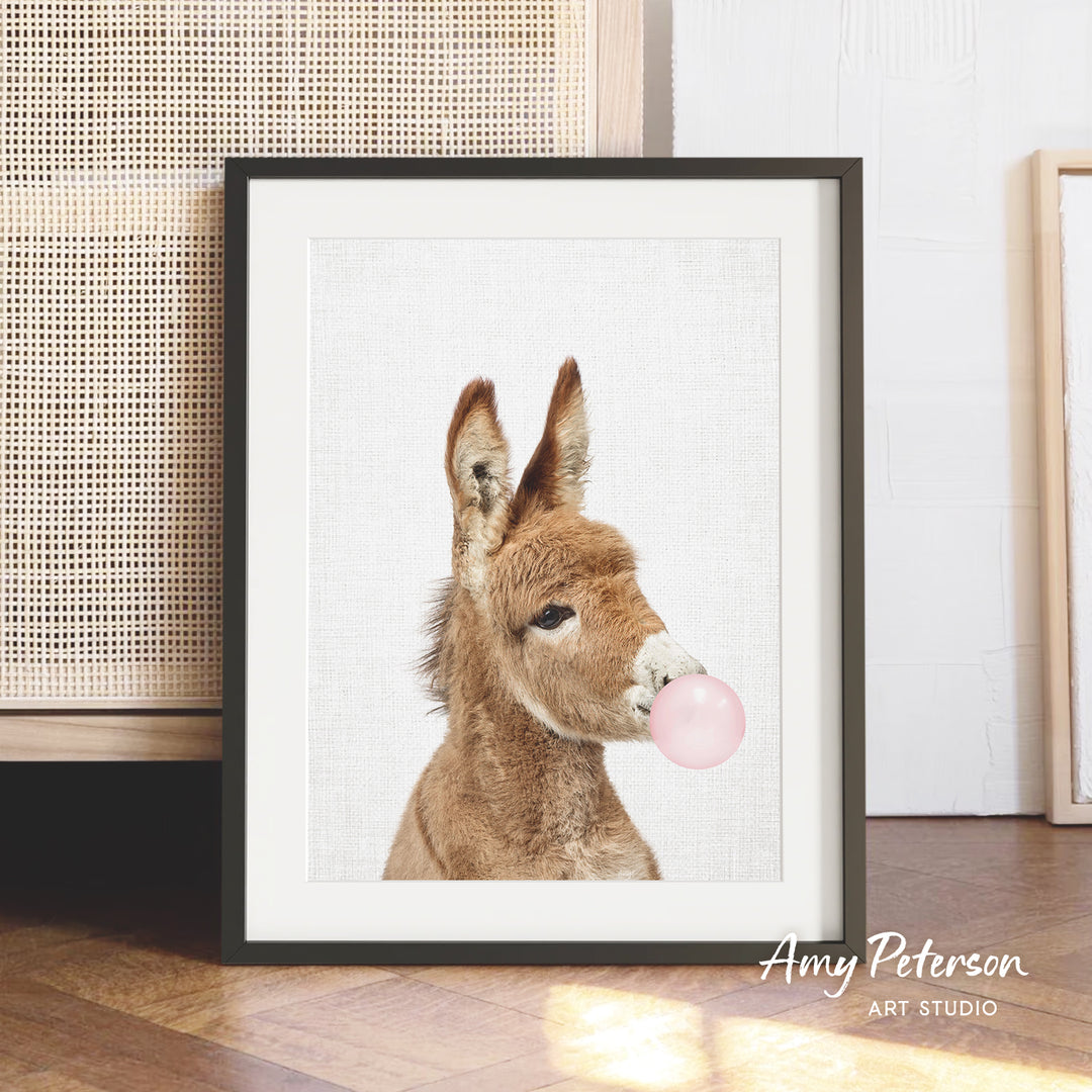 A framed photograph of a donkey blowing a bubble with a pink bubble gum bubble.