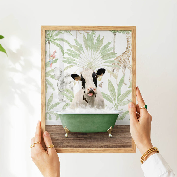 A person's hands are holding a framed picture of a cow sitting in a green bathtub, surrounded by tropical foliage.