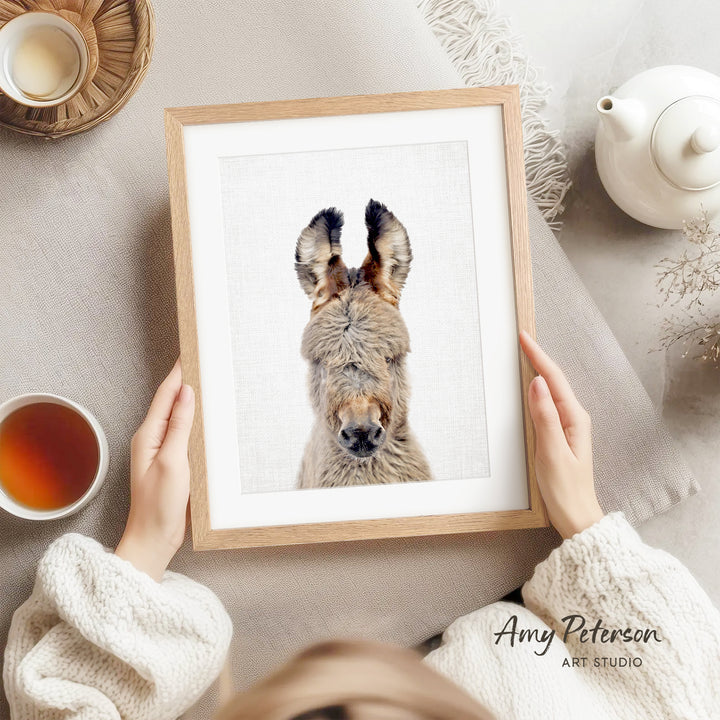 A person is holding a framed picture of a donkey with its head turned to the side, while sitting on a couch with a cup of tea nearby.