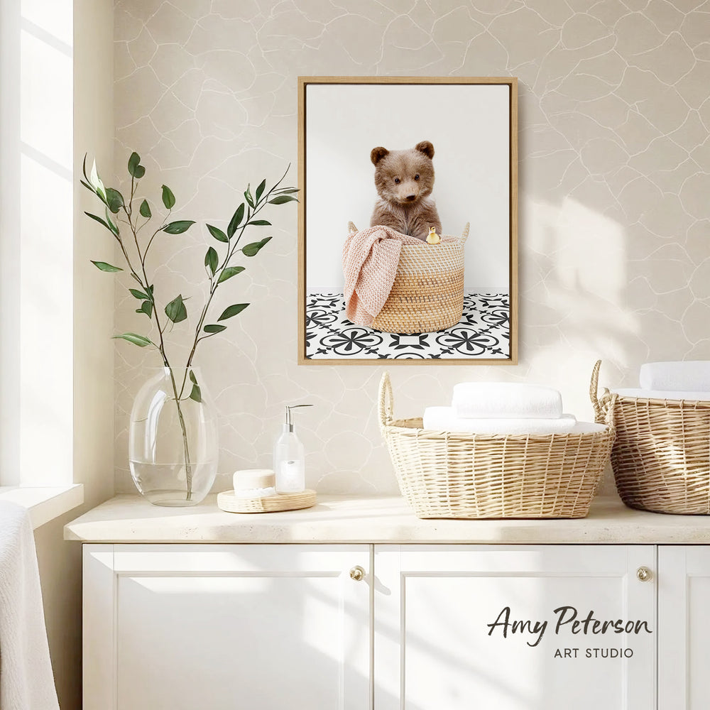 a cozy bathroom scene with a framed photo of a cute bear sitting in a woven basket, placed on a wall above a white cabinet. The cabinet has two baskets on top, one of which is filled with white towels. There is also a vase with green leaves and a bottle of soap on the countertop. The overall scene has a warm and inviting atmosphere.