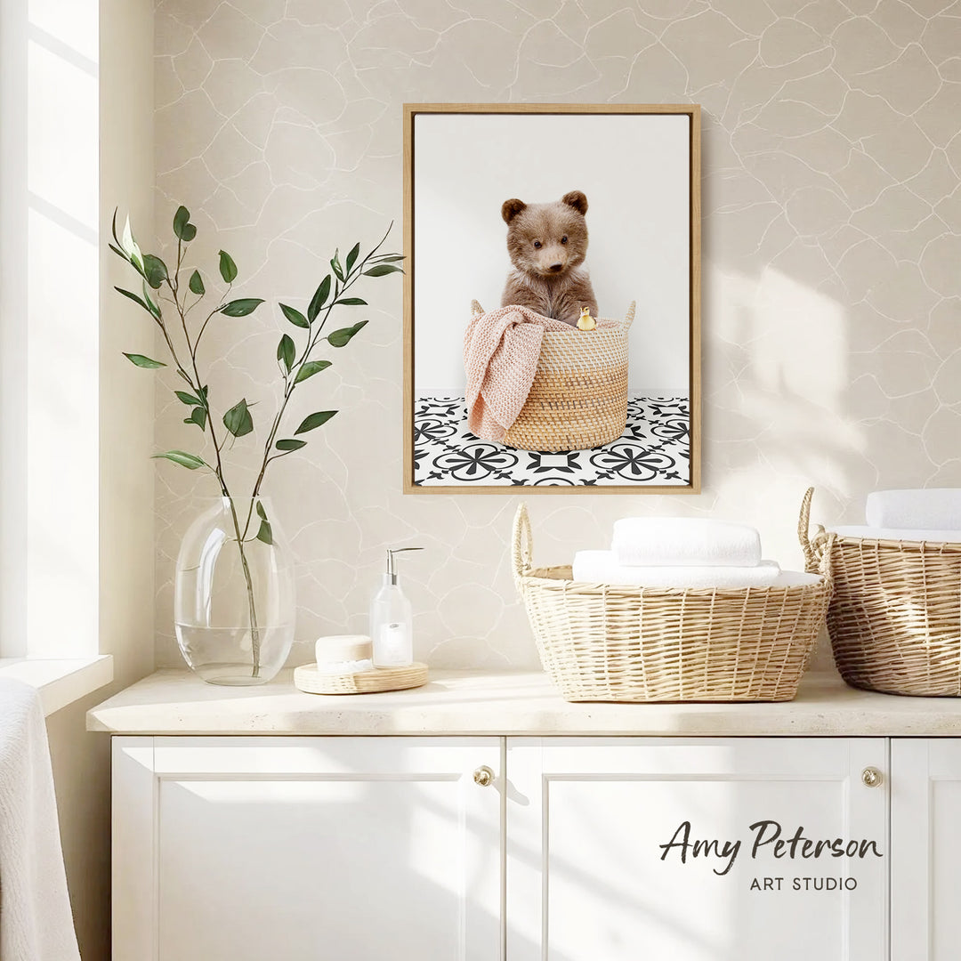 a cozy bathroom scene with a framed photo of a cute bear sitting in a woven basket, placed on a wall above a white cabinet. The cabinet has two baskets on top, one of which is filled with white towels. There is also a vase with green leaves and a bottle of soap on the countertop. The overall scene has a warm and inviting atmosphere.