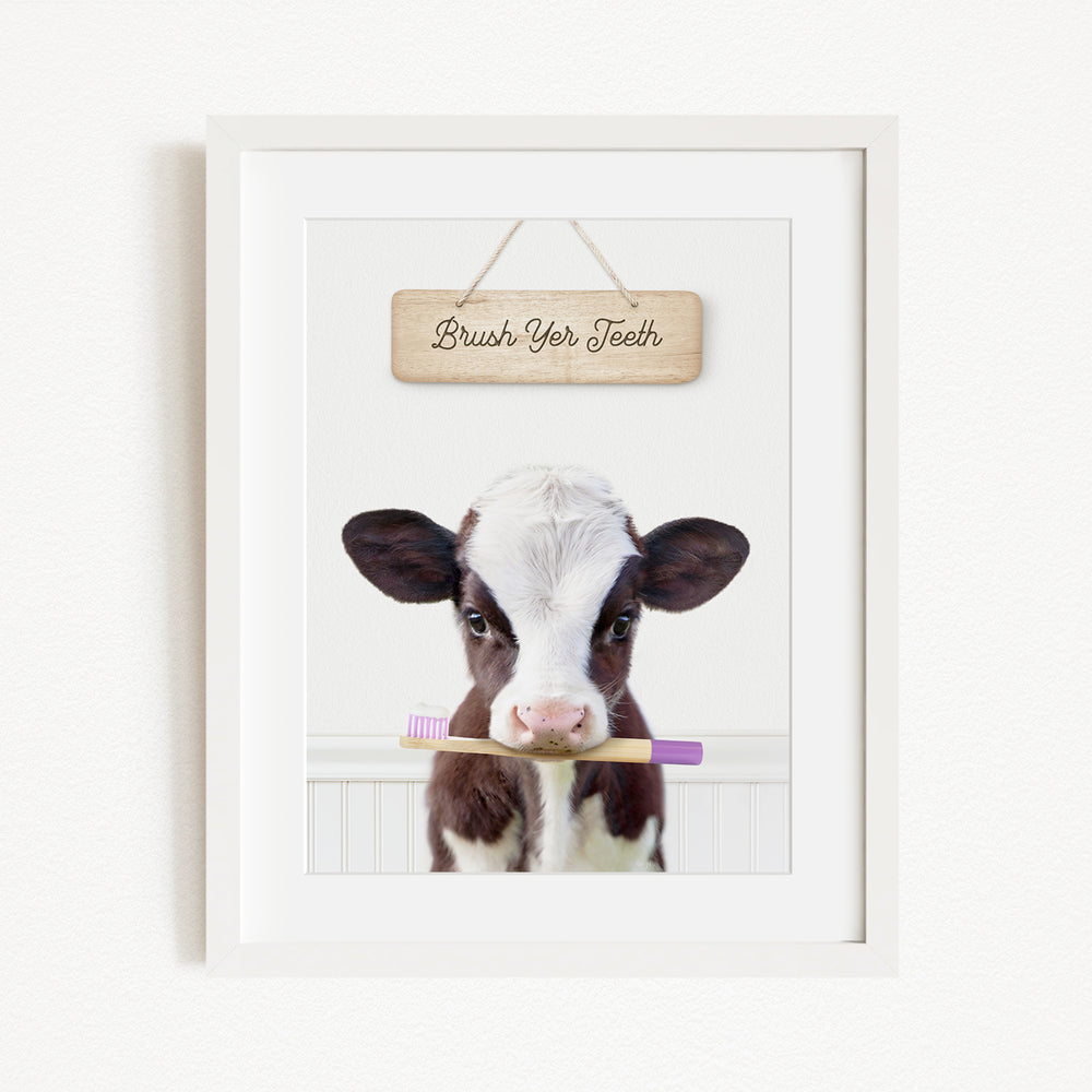 A framed image of a cow holding a purple toothbrush in its mouth, with a wooden sign above that says "Brush Your Teeth".