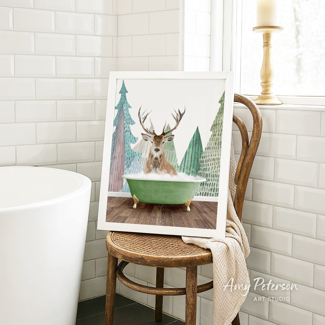A framed image of a deer sitting in a bathtub, surrounded by Christmas trees, is displayed on a wooden chair in a bathroom setting.