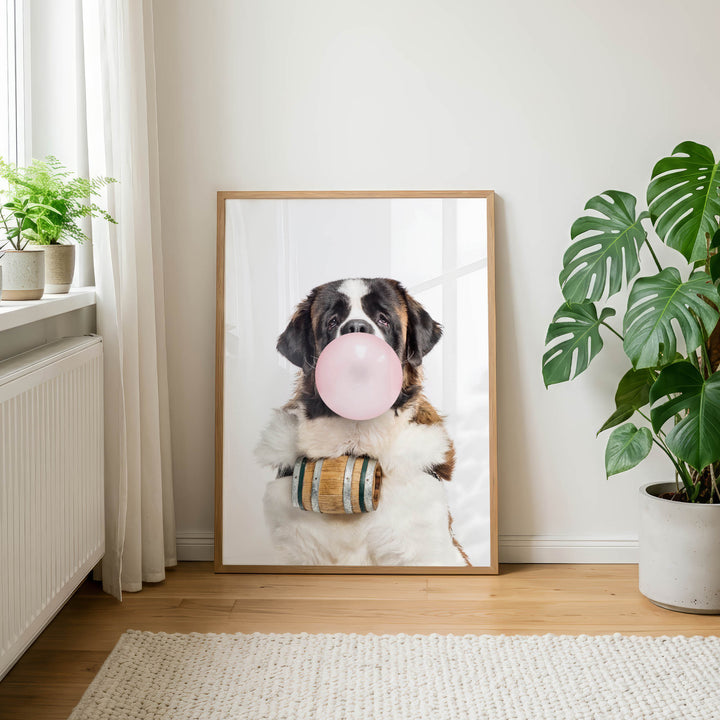 A framed photograph of a dog blowing a bubble with a pink bubble gum bubble, wearing a wooden barrel around its neck, and sitting on a wooden floor in a room with a window and potted plants.