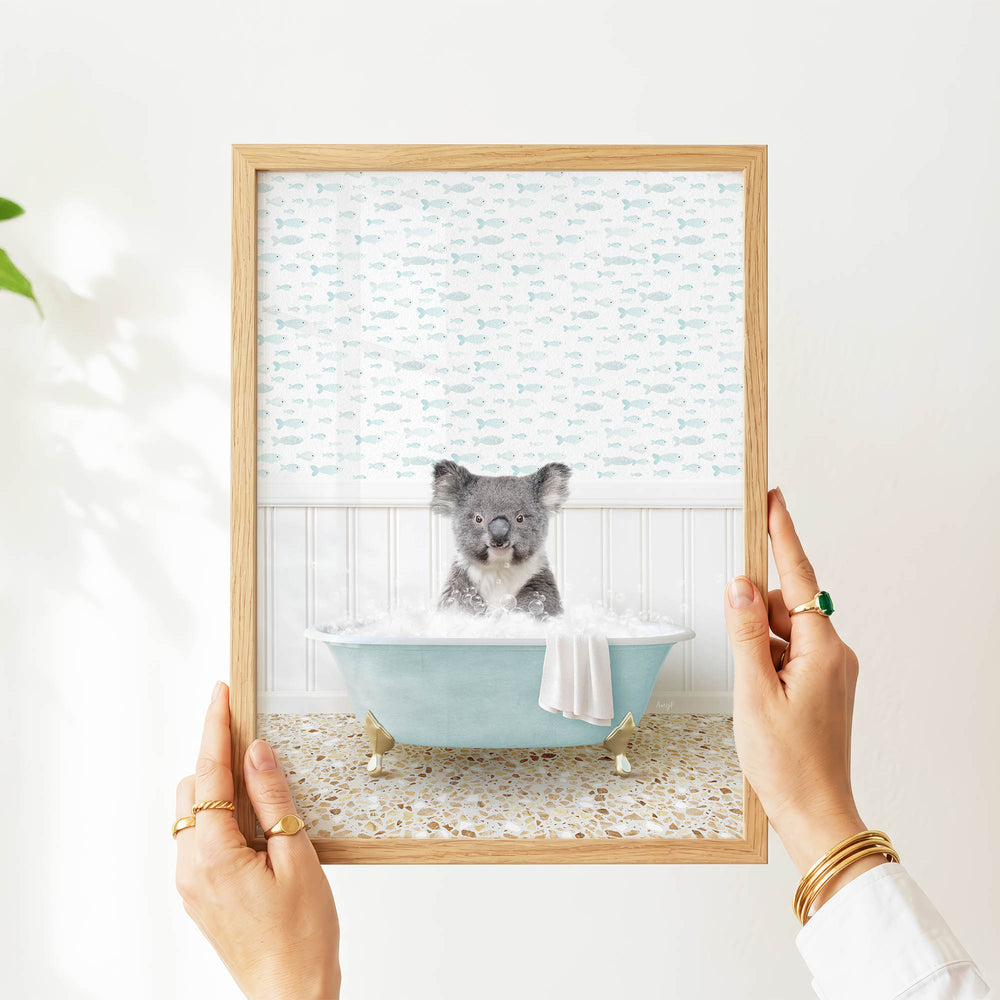 A person's hands are holding a framed picture of a koala bear sitting in a bathtub filled with bubbles, surrounded by a pattern of fish.
