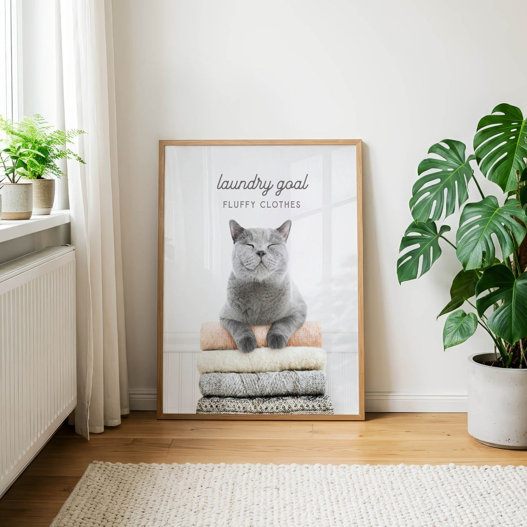 A framed poster with a cat sitting on a pile of clothes, with the text "laundry goal" and "FLUFFY CLOTHES" displayed on it.