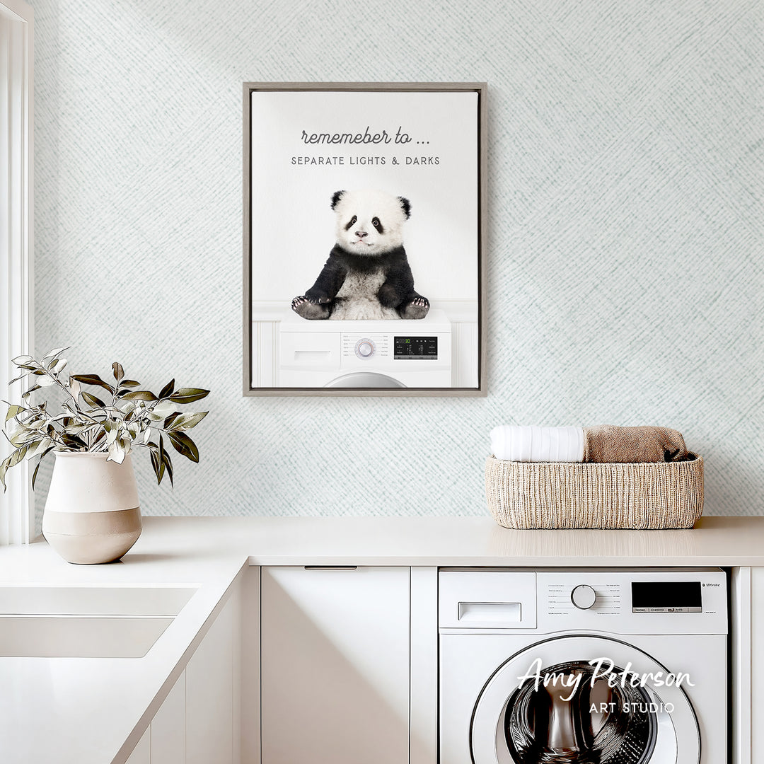 a panda bear sitting on a washing machine, with a framed poster of the panda bear above it. The poster has the text "Remember to..." and "Separate Lights & Darks". The panda bear is sitting on a white washing machine, which is placed on a white countertop. There is also a basket of towels and a plant in the image.