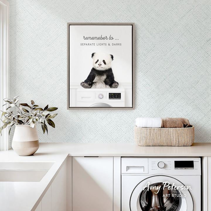 a panda bear sitting on a washing machine, with a framed poster of the panda bear above it. The poster has the text "Remember to..." and "Separate Lights & Darks". The panda bear is sitting on a white washing machine, which is placed on a white countertop. There is also a basket of towels and a plant in the image.