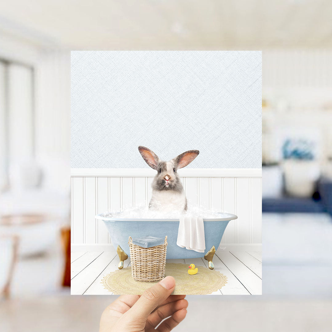 A hand holds up a framed image of a rabbit sitting in a bathtub filled with bubbles, surrounded by a yellow rubber duck and a white towel.