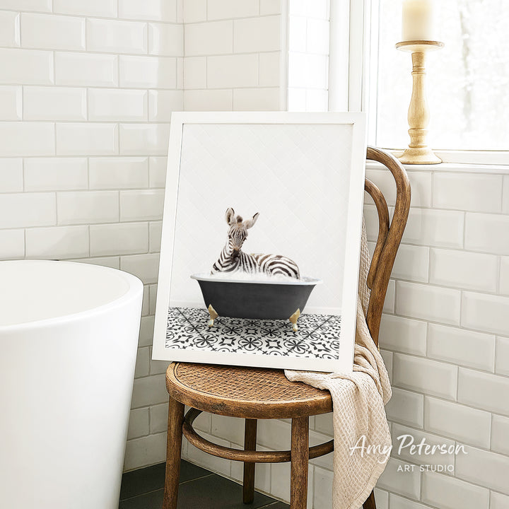 A framed image of a zebra sitting in a bathtub is displayed on a wooden chair in a bathroom setting.
