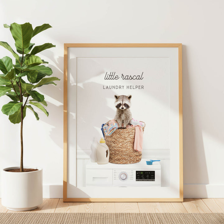A framed image of a raccoon sitting on a laundry basket, with various laundry-related items such as a detergent bottle and a washing machine visible in the background.