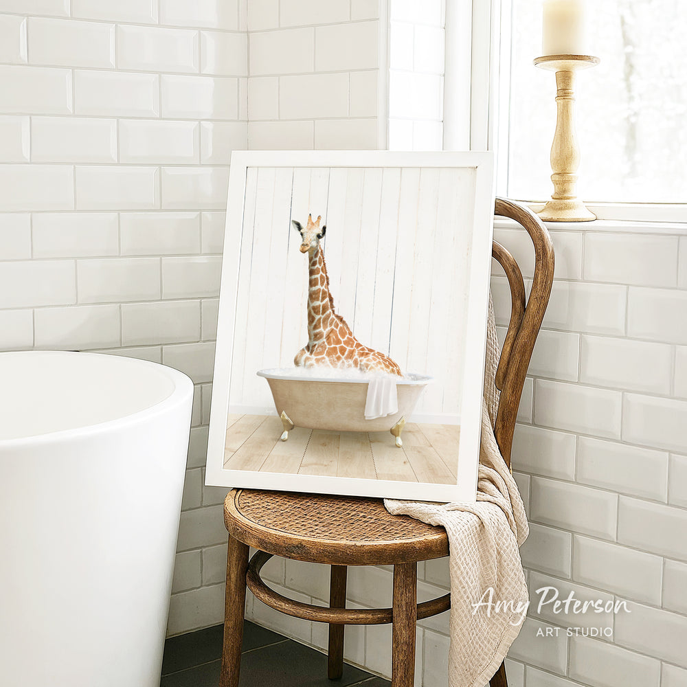 A framed image of a giraffe sitting in a bathtub is displayed on a wooden chair in a bathroom setting.