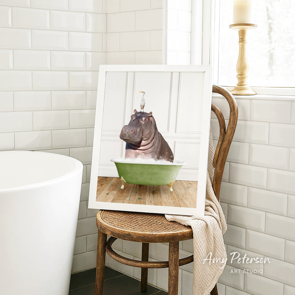 A framed image of a hippopotamus sitting in a green bathtub, with a bird perched on the hippopotamus' head, is displayed on a wooden chair in a bathroom setting.