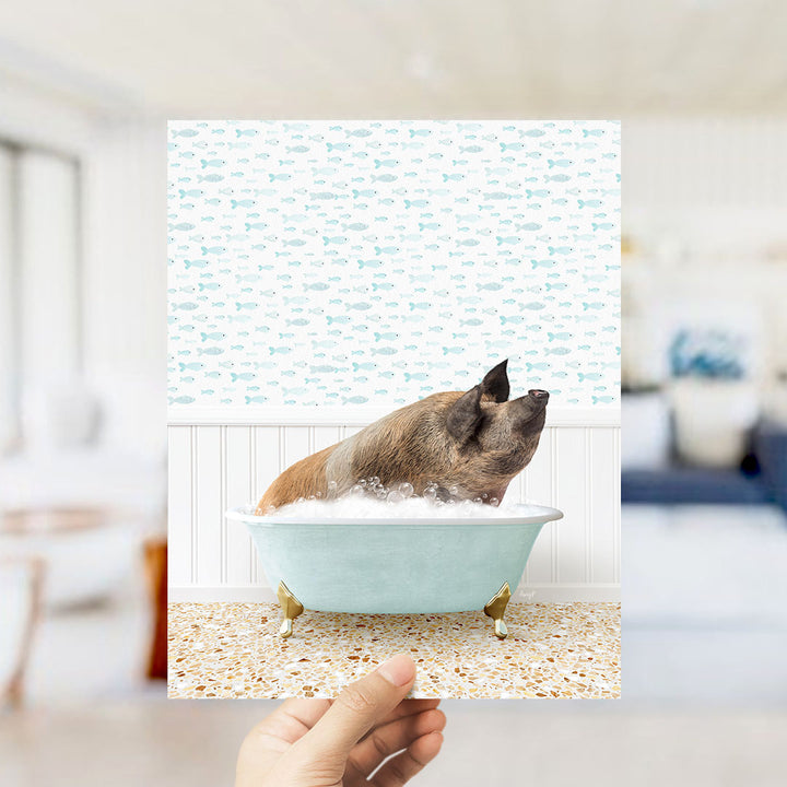 A hand holds up a framed image of a pig sitting in a bathtub filled with bubbles, surrounded by a pattern of small fish.