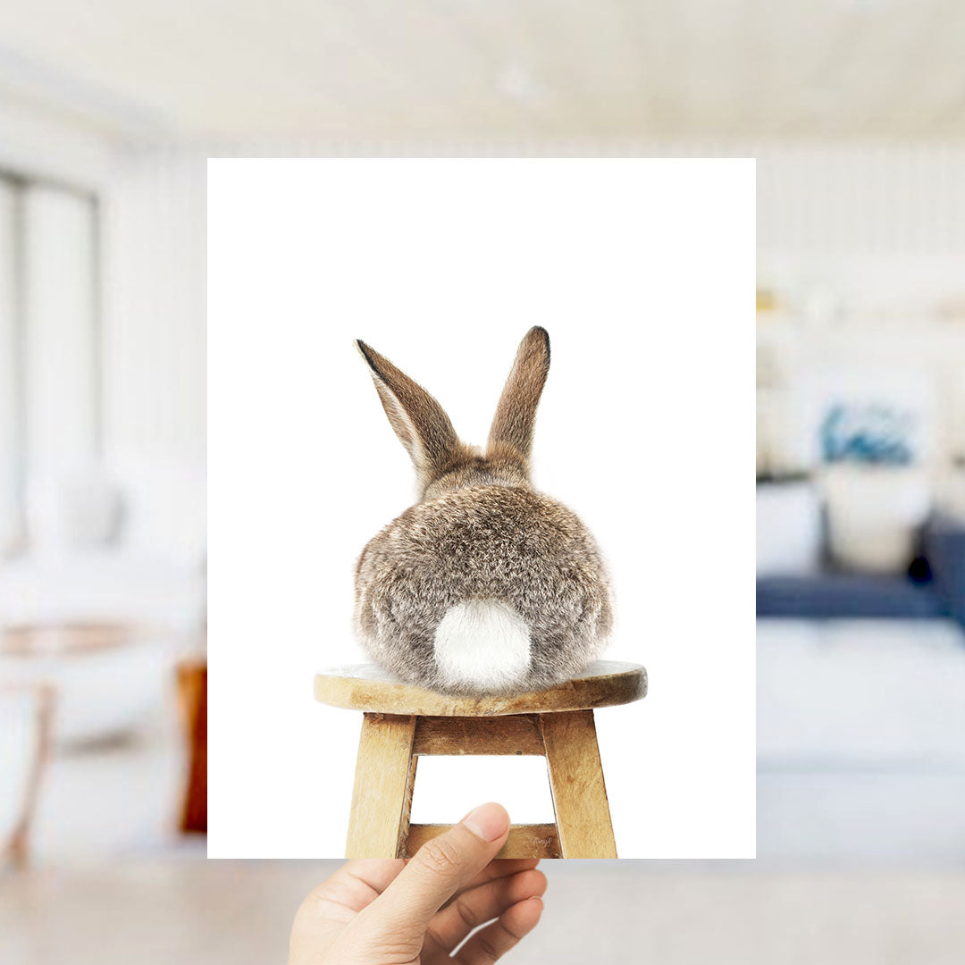A person's hand is holding up a picture of a rabbit sitting on a wooden stool.