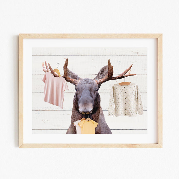 A framed collage of a moose holding clothes, including a pink shirt and a white sweater with polka dots, against a white background.