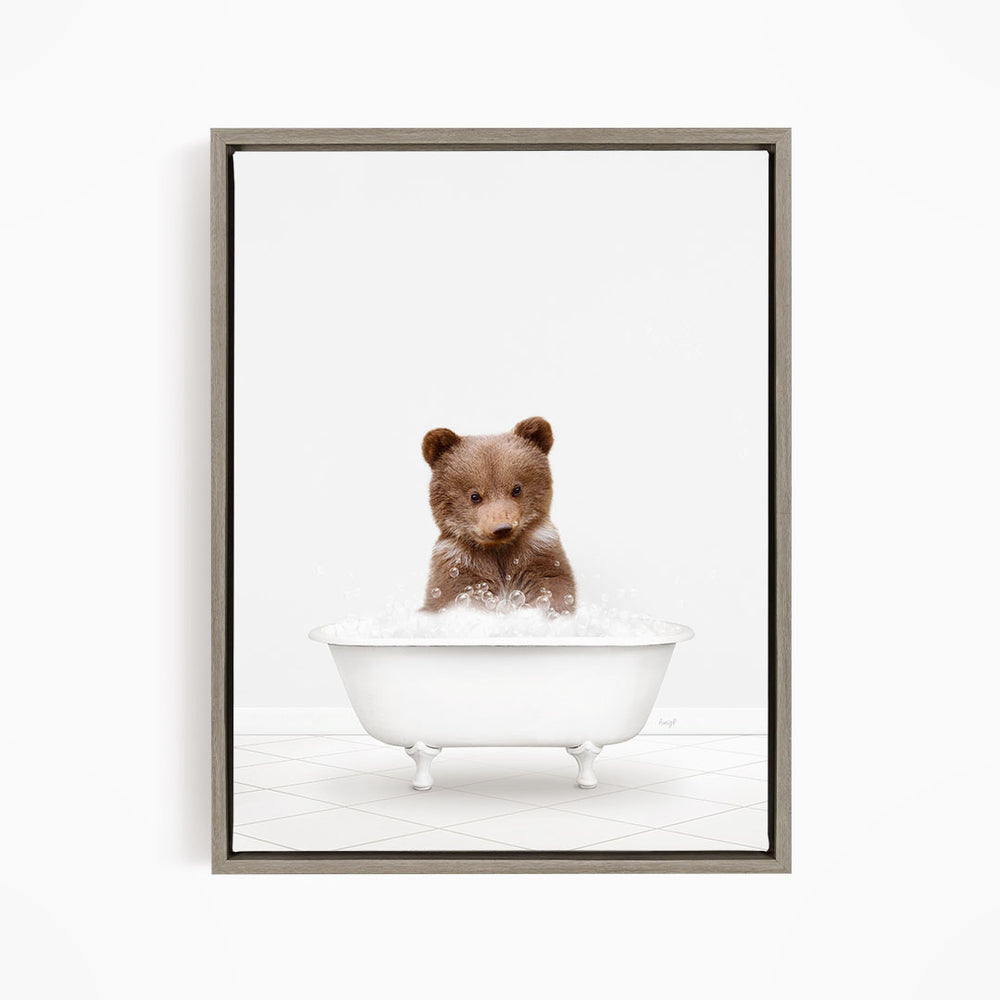 A cute brown bear cub sitting in a white bathtub filled with bubbles, looking directly at the camera.