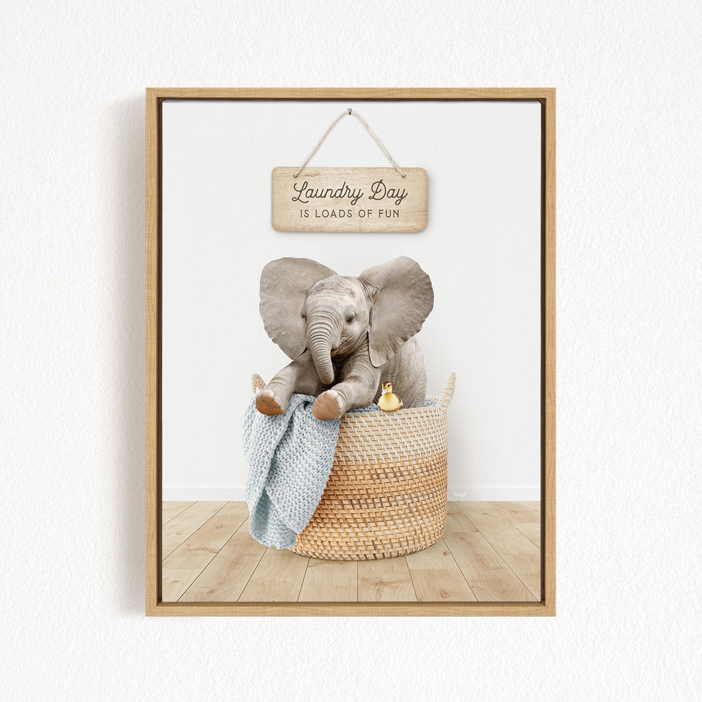 A framed image of a baby elephant sitting in a woven basket, with a sign above that says "Laundry Day is loads of fun".