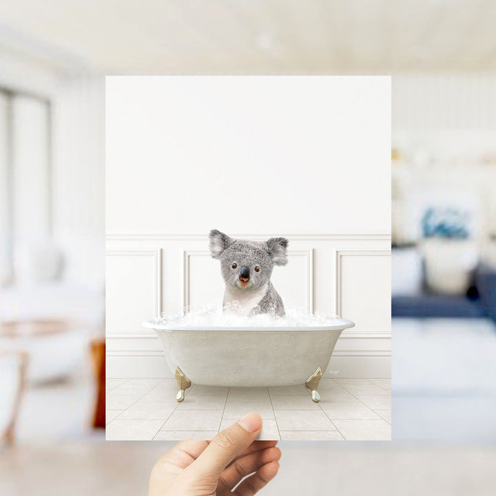 A hand holds up a photograph of a koala bear sitting in a bathtub filled with water, creating a playful and whimsical scene.