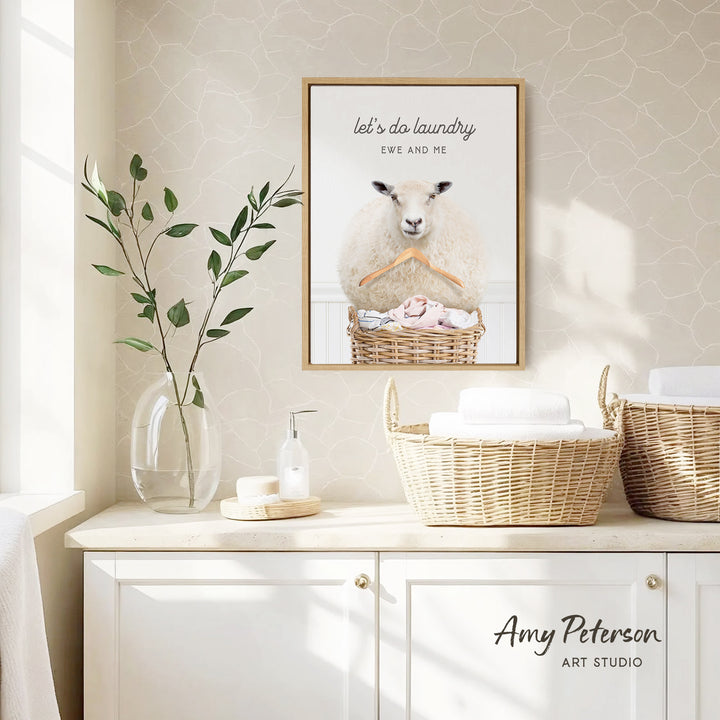 a framed picture of a sheep hanging on a wall, with the text "let's do laundry EWS AND ME" above it. The wall has a beige pattern, and there are baskets and a vase with greenery on the counter.