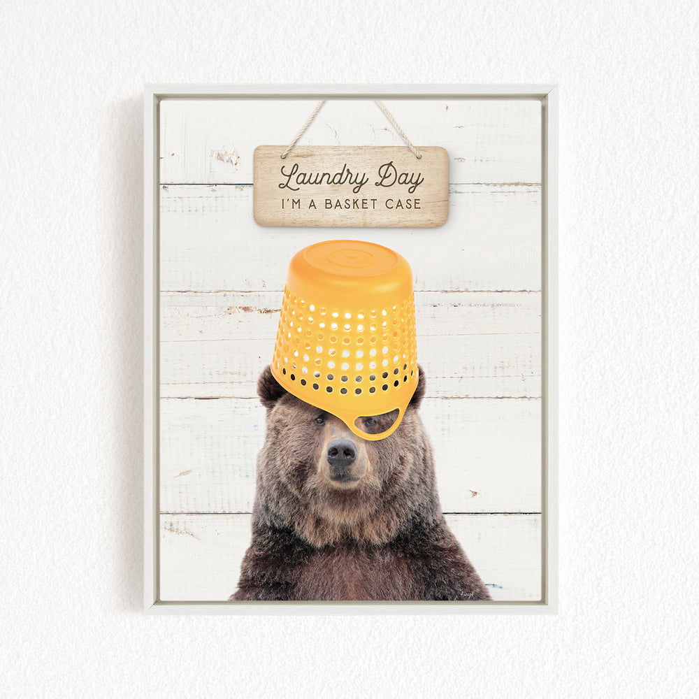 A framed image of a bear wearing a yellow plastic basket on its head, with a wooden sign above it that says "Laundry Day I'm a basket case".