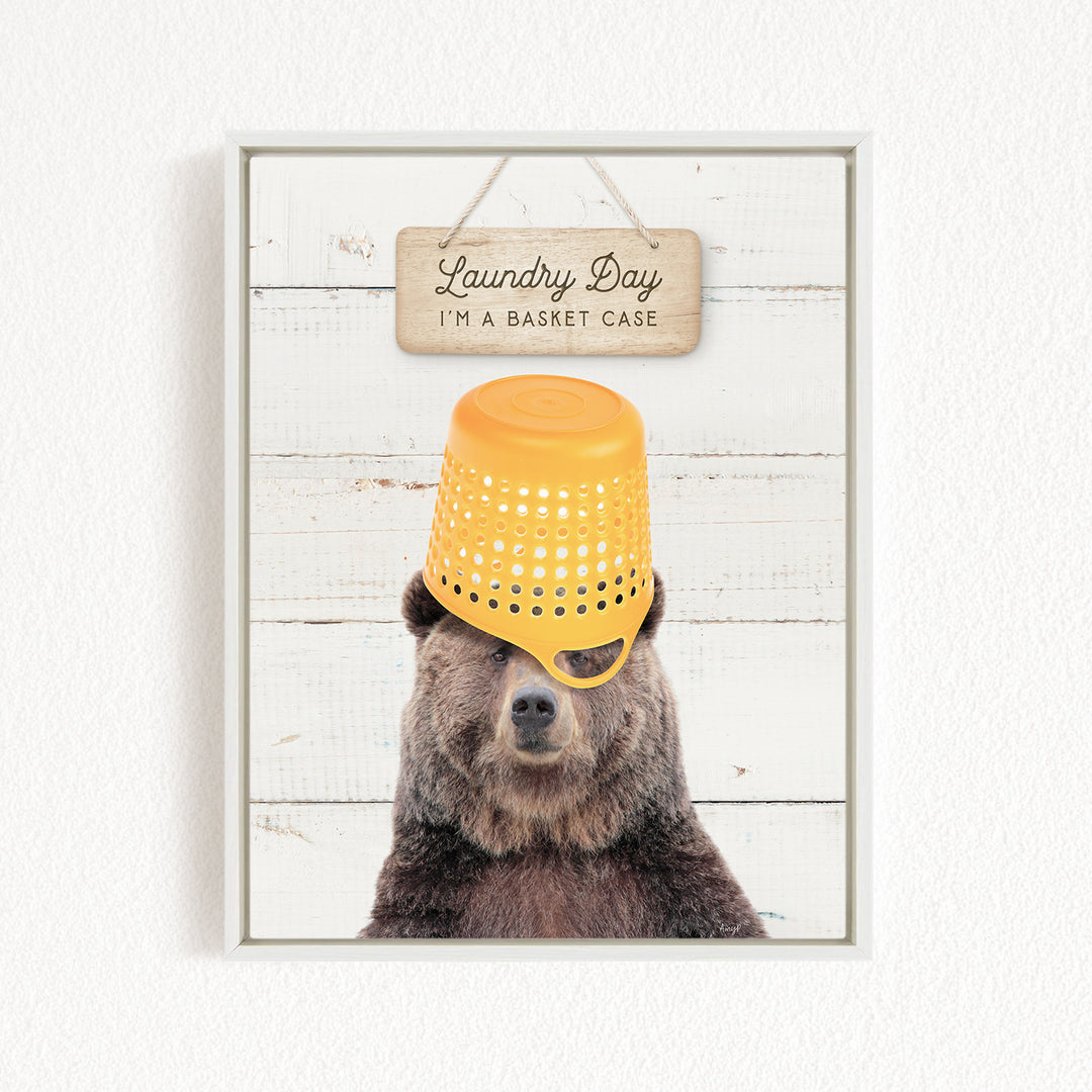 A framed image of a bear wearing a yellow plastic basket on its head, with a wooden sign above it that says "Laundry Day I'm a basket case".