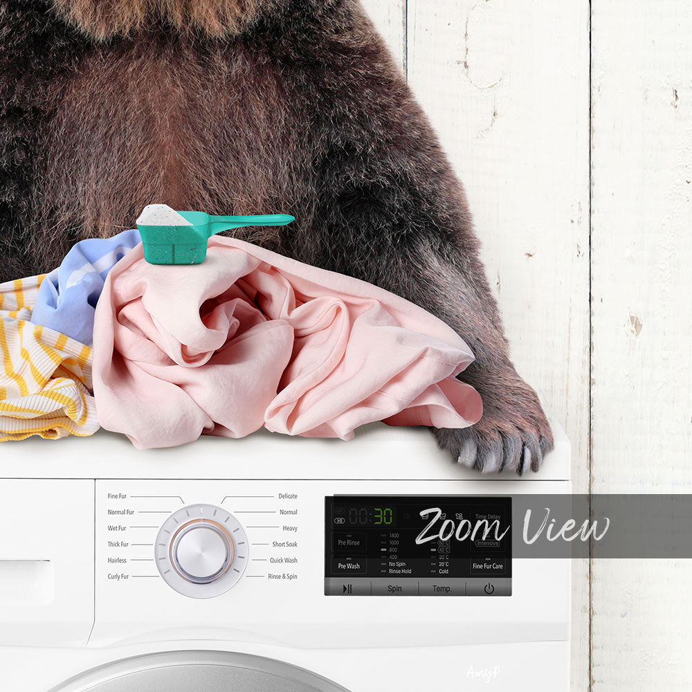A brown bear is sitting on top of a white washing machine, with laundry items such as clothes and towels nearby.