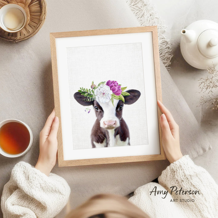 A person is holding a framed picture of a cow with a floral headpiece, surrounded by various objects such as a teapot, a cup, and a bowl.