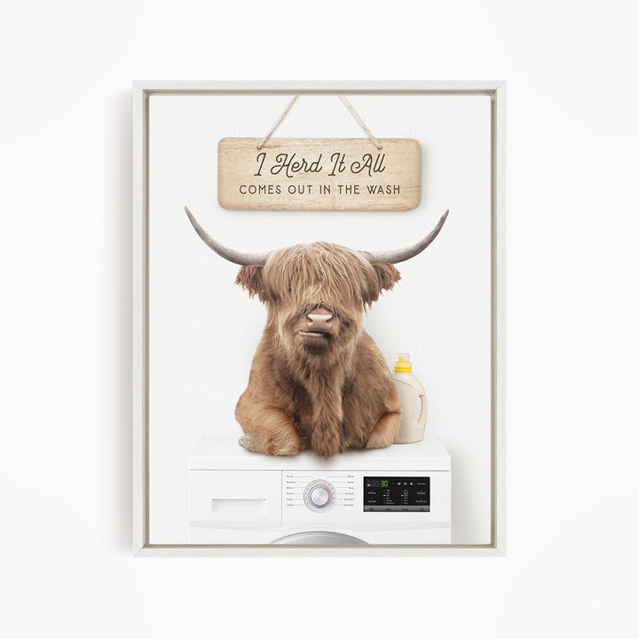 A brown cow with long horns sits on top of a white washing machine, with a wooden sign above it that says "I Herd It All Comes out in the Wash".