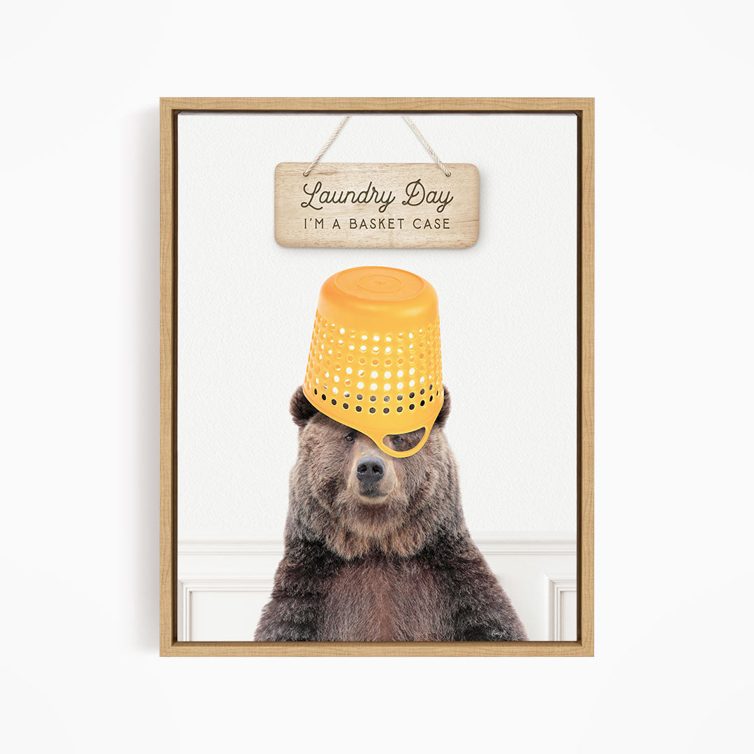 A framed image of a bear wearing a yellow plastic basket on its head, with a wooden sign above it that says "Laundry Day I'm a basket case".