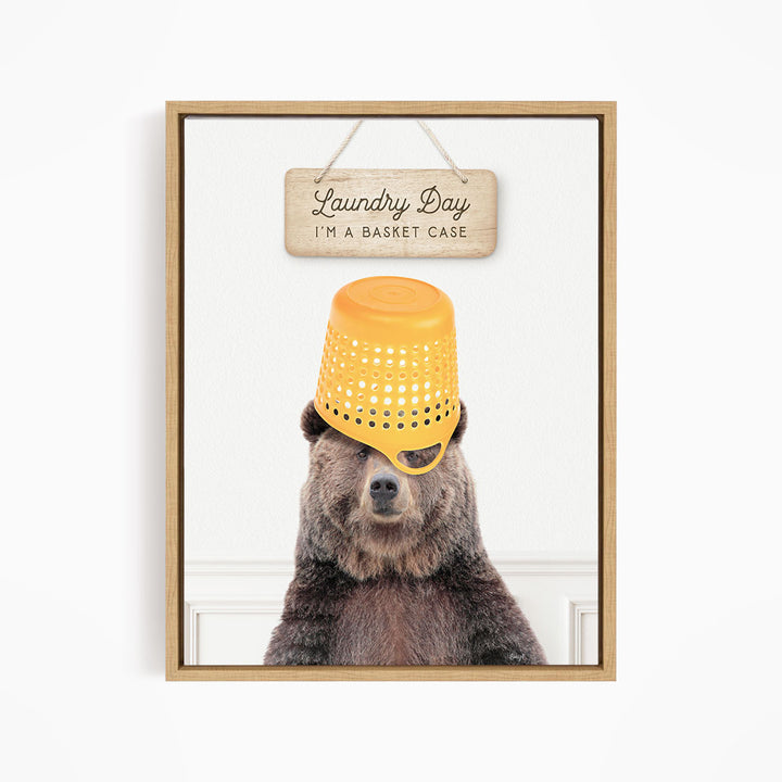 A framed image of a bear wearing a yellow plastic basket on its head, with a wooden sign above it that says "Laundry Day I'm a basket case".