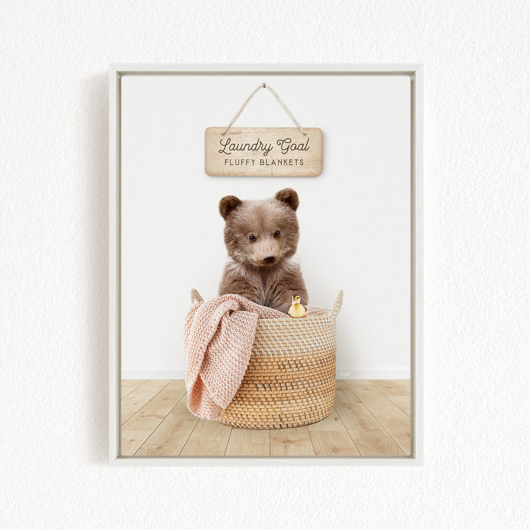 A framed image of a brown teddy bear sitting in a woven laundry basket, with a wooden sign above that reads "Laundry Goal Fluffy Blankets".